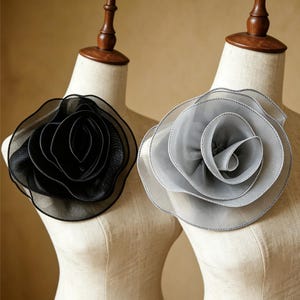 May include: Two large flower brooches made of layered, sheer, translucent fabric. One brooch is solid black, and the other is light gray. Each flower features multiple ruffled petals with visible stitching along the edges, displayed on two beige mannequins.