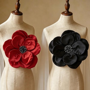 May include: Two fabric flower brooches displayed on beige mannequins. One flower is deep red and the other is black. Both feature layered, rounded satin-like petals and a central cluster of small, white, bead-like accents at the core.