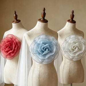 May include: Three beige dress forms each displaying a large, layered fabric flower on the chest. The flowers are red, light blue, and white, featuring delicate, translucent petals with dark stitched edges and sheer fabric draping from the shoulders.