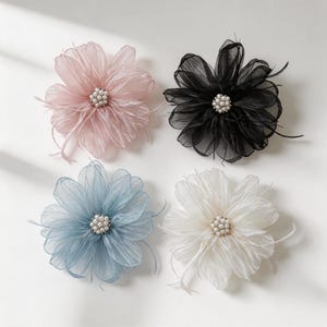 Puede incluir: Cuatro accesorios para el cabello en forma de flor hechos de pétalos de tela transparente en capas, en colores rosa claro, negro, azul polvoriento y blanco roto. Cada flor tiene un centro de pequeñas perlas blancas y detalles de plumas.