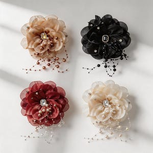 Puede incluir: Cuatro broches florales en beige, negro, rojo oscuro y crema. Cada broche presenta pétalos de tela superpuestos, un estrás rectangular central y delicados hilos colgantes de cuentas y perlas a juego que salen del centro.
