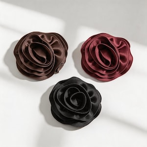 May include: Three fabric flower accessories in dark brown, deep burgundy, and black. Each piece features layered, ruffled satin-like petals arranged in a circular, rose-like shape with a smooth, reflective finish and soft, curved edges.