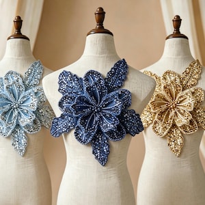 Può includere: Tre applicazioni floreali in tessuto su manichini sartoriali beige. Quella a sinistra è azzurra, quella centrale è blu navy scuro e quella a destra è dorata. Ogni fiore presenta petali stratificati ricoperti di paillettes e perline luccicanti.