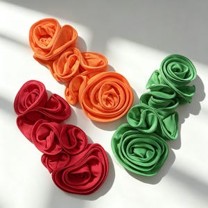 May include: Three fabric floral appliques in bright orange, deep red, and vibrant green. Each piece features multiple layered, spiraled fabric petals arranged in a long, flowing shape with finished edges, creating a dimensional, textured appearance.