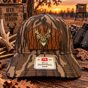 May include: A camouflage baseball cap featuring a brown leather-like patch with a deer head illustration. The cap has a white rectangular sticker on the brim with the text Est. 1970, R, It's a Richardson, OSFM. The fabric has a brown and black bark pattern.