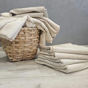 May include: A woven basket overflowing with folded beige linens, next to a stack of folded linens. The basket is light brown, and the linens are a natural, neutral color. The background is a gray wall and light wood floor.