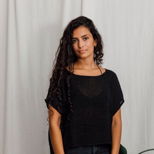 May include: Black, short-sleeved top with a relaxed fit and a boat neck. The top is worn with dark wash jeans. The model has long, dark curly hair and is standing in front of a white curtain.