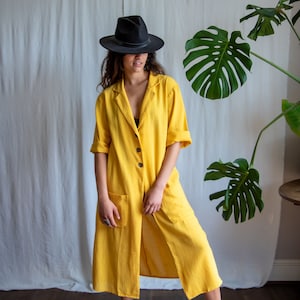 May include: A woman wearing a yellow linen dress with a black hat and standing on a wooden floor. A large green plant is in the background.
