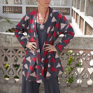 May include: A woman wearing a long-sleeved, dark grey and red patterned jacket with a silver necklace and earrings. The jacket has a geometric pattern of triangles.