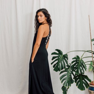 Puede incluir: Una mujer con un vestido maxi negro con un detalle blanco en forma de cruz en la espalda. Está de pie frente a una pared blanca con una gran planta verde en el fondo.
