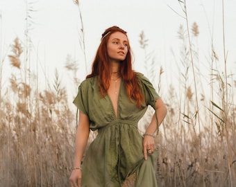 May include: A woman with long, red hair wearing a green maxi dress stands in a field of tall, dried grasses. She is looking off to the side and has a relaxed expression on her face.