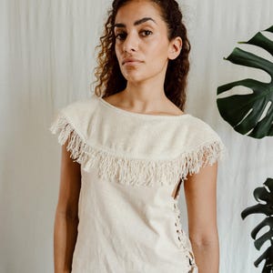 Op de afbeelding: Een crèmekleurige mouwloze top met een gefranjerde halslijn. De top heeft een eenvoudig ontwerp met een ronde hals en veters aan de zijkant. Het model draagt de top met een zwart-wit patroon rok.