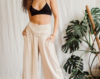 May include: A woman wearing a black halter top and loose-fitting, light beige pants with a knotted waistband. She is standing in front of a white wall with a large green plant in the background.