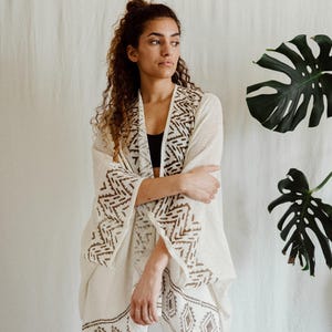 May include: A cream-coloured kimono-style cardigan with brown geometric embroidery on the sleeves and hem. The cardigan is worn over a black top and white trousers. A large green leaf is visible in the background.