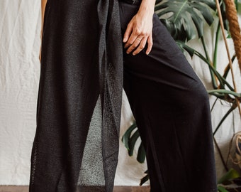 May include: Black wide-leg pants with a long, sheer overlay tied at the waist. The overlay is made of a fine mesh fabric.
