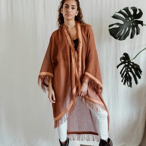 May include: A long, open-front brown kimono with fringed edges and decorative trim. The kimono is worn over a black top and white pants. Black cowboy boots complete the look. The background is a neutral-colored fabric.