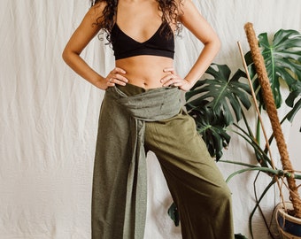 May include: A woman wearing a black bra and green pants with a wrap-around tie. The pants are wide-legged and flowy. The woman is standing in front of a white wall with a plant in the background.