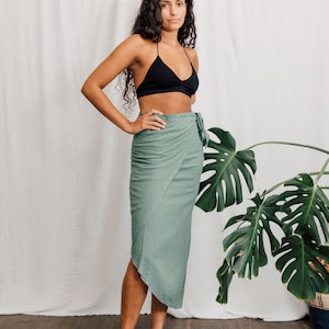 May include: A woman wearing a black bra and a sage green wrap skirt. She is standing in front of a white wall with a large green plant in the background.