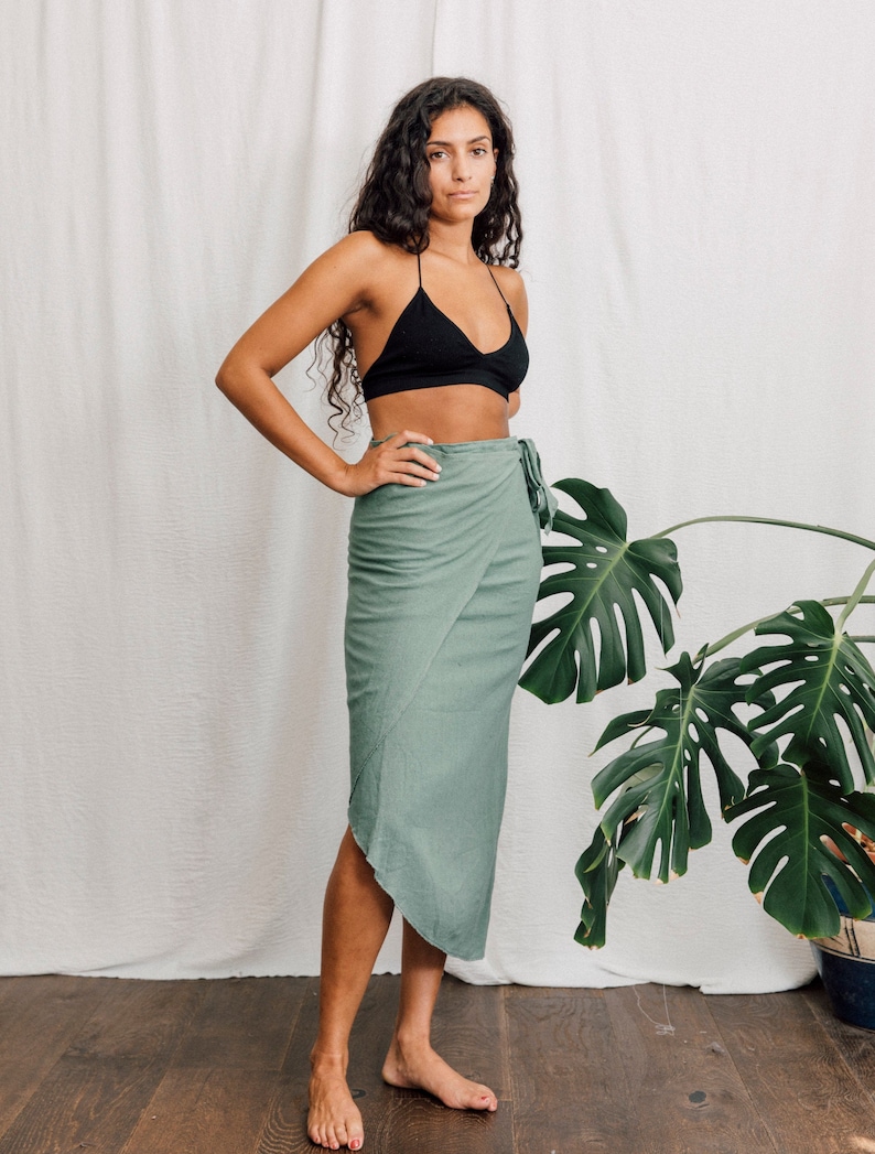 May include: A woman wearing a black bra and a sage green wrap skirt. She is standing in front of a white wall with a large green plant in the background.