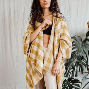 May include: A woman wearing a yellow and white checked kimono with a black bra underneath. She is wearing beige trousers and has long curly brown hair.