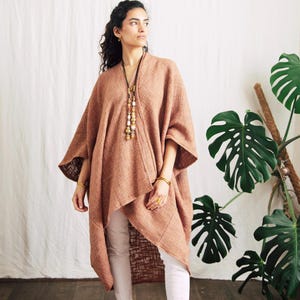 May include: A woman in a rust-coloured poncho-style top with a draped front and wide sleeves, paired with white trousers. A long necklace with beads and gold bracelets complete the look. A large-leafed plant is in the background.