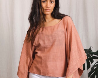 Puede incluir: Una mujer lleva una blusa de lino holgada de color salmón con mangas largas y cuello redondo. Lleva pantalones vaqueros blancos y tiene el pelo largo y oscuro.