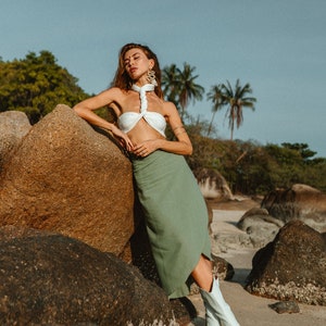 May include: A woman wearing a white halter top and a green skirt stands on a beach with large rocks. She is wearing white boots and has her eyes closed.