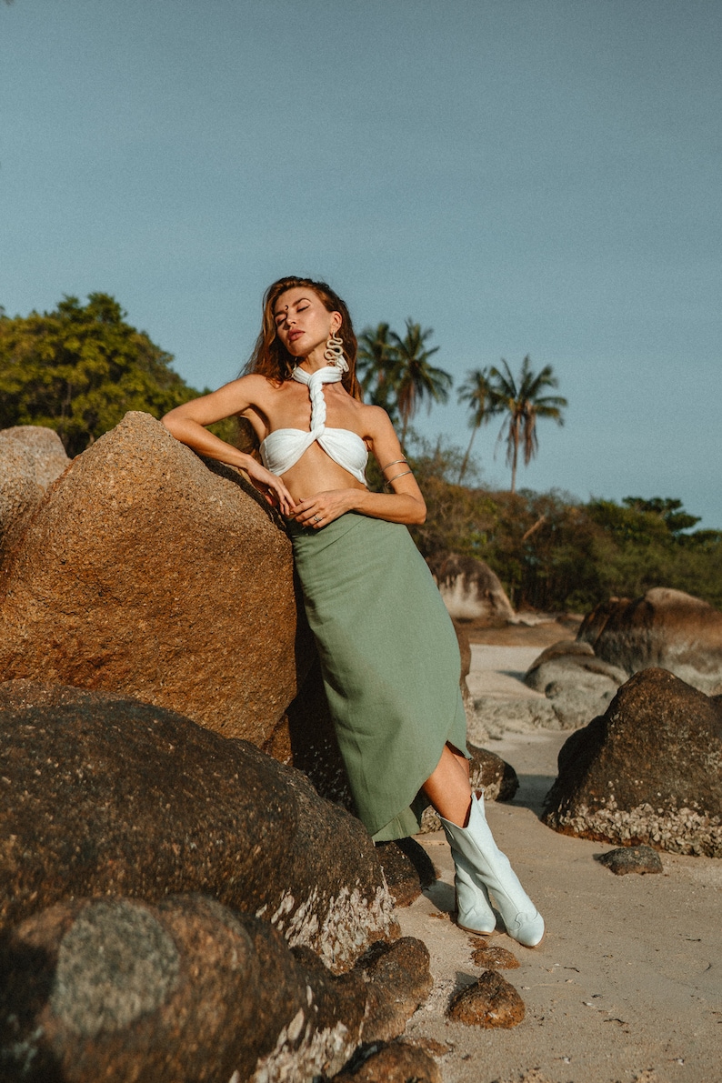 May include: A woman wearing a white halter top and a green skirt stands on a beach with large rocks. She is wearing white boots and has her eyes closed.