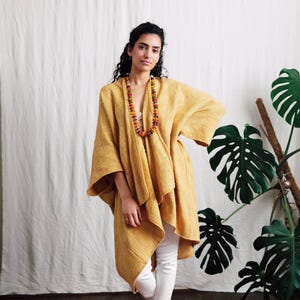 May include: A mustard yellow open-front kimono-style wrap with wide sleeves and a textured fabric finish. The garment is paired with white trousers, a long multi-coloured beaded necklace, and a small turquoise-patterned anklet on the left ankle.