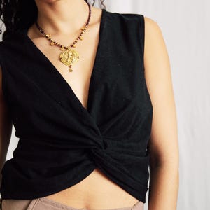 May include: A black sleeveless top with a front twist detail at the hem, paired with a gold-toned necklace featuring a circular pendant and dark red beads. A gold-toned chain belt with circular accents is worn over light brown trousers with buttons.