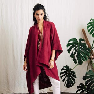May include: A burgundy open-front kimono jacket with wide sleeves and a draped design. The model is wearing white trousers and gold jewellery. A large-leafed plant is in the background.
