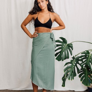 May include: A woman wearing a black halter top and a sage green wrap skirt. She is standing in front of a white backdrop with a large green plant to the right.