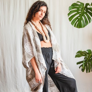 May include: A woman wearing a white and grey crochet kimono with black trousers and a shell necklace. She is standing in front of a white wall with a green plant in the background.
