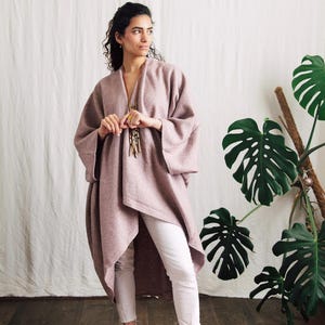 May include: A mauve-colored, open-front coat with wide sleeves. The coat is paired with white pants. A long necklace with multiple strands hangs from the neck. The background includes a large-leafed plant and a white backdrop.