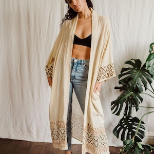 Op de afbeelding: Een lange, crèmekleurige kimono met bruine en groene geometrische patronen op de mouwen en zoom. De kimono wordt gedragen over een zwarte bh en een blauwe jeans.