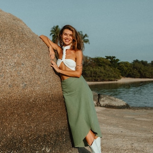 May include: A woman wearing a white halter top and a green skirt stands next to a large rock on a sandy beach. She is smiling and looking at the camera. She is wearing white cowboy boots.