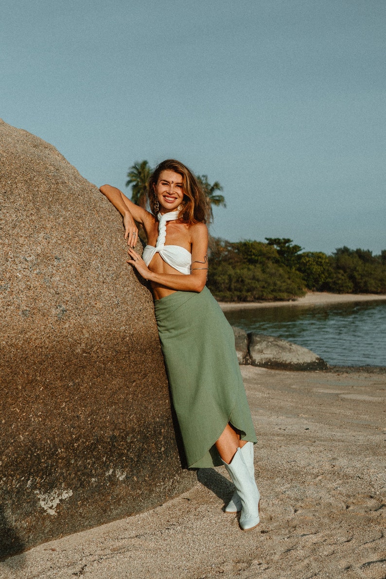 May include: A woman wearing a white halter top and a green skirt stands next to a large rock on a sandy beach. She is smiling and looking at the camera. She is wearing white cowboy boots.