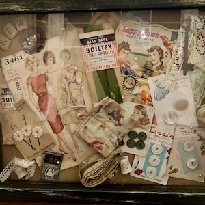 May include: A framed shadow box filled with vintage sewing notions. Includes dress patterns, bias tape, buttons, lace, and a measuring tape. The word "SEW" is spelled out with individual letter tiles. The box has a distressed black frame.