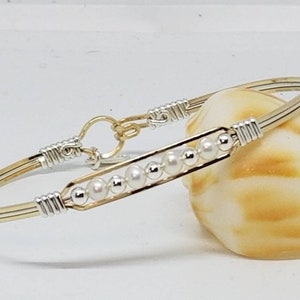 Puede incluir: Un brazalete de alambre envuelto en oro y plata con una fila de perlas blancas y cuentas de plata.