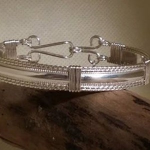 May include: A silver bracelet with a braided design and a smooth, flat center section. The bracelet has a clasp closure.