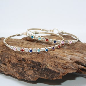 May include: Two silver wire wrapped bracelets with colorful gemstones. The bracelets are on a piece of driftwood.