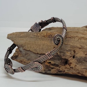 May include: A copper and silver wire bracelet with a swirl design. The bracelet is on a piece of driftwood.