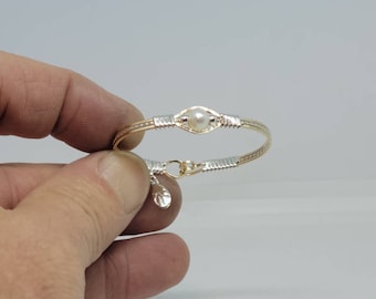 Puede incluir: Un brazalete de alambre envuelto en oro y plata con un solo dije de perla blanca. El brazalete tiene un pequeño dije que cuelga.
