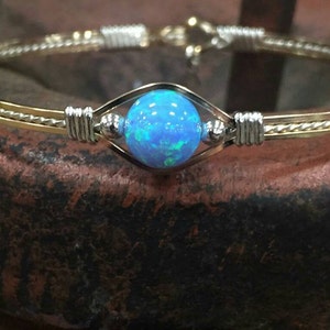 May include: A gold and silver bangle bracelet with a central blue opal stone. The bracelet features a round blue opal gemstone set in a diamond-shaped silver setting. The bracelet is made of gold and silver metal.