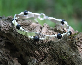 May include: A silver wire wrapped bracelet with black beads. The bracelet is on a piece of wood.