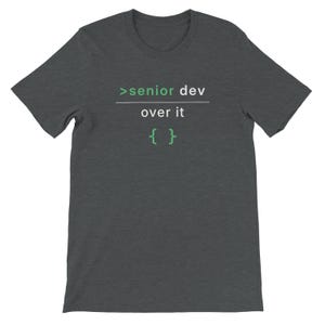 May include: A dark grey t-shirt featuring the text ">senior dev over it { }" in green and white. This short-sleeved crew neck top is designed for casual wear. The text is centrally placed on the chest.