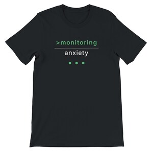 Può includere: T-shirt nera con la scritta ">monitoring anxiety" in verde e bianco. Il testo è centrato sul davanti. Sotto la parola "anxiety" ci sono tre punti verdi.
