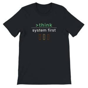 May include: Black t-shirt featuring the text ">think system first" in white and green. Below the text are three vertical rectangles in shades of brown and orange. The shirt has a crew neck and short sleeves.