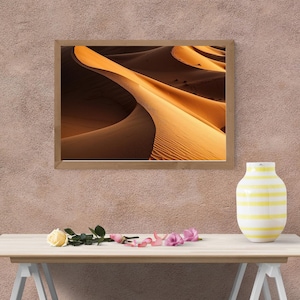 Könnte beinhalten: Ein gerahmter Druck von Sanddünen in der Wüste. Das Bild zeigt warme Töne von Beige und Braun, mit Schatten und Lichtern, die einen strukturierten Effekt erzeugen. Der Druck wird über einem Tisch mit einer Vase und Blumen angezeigt.
