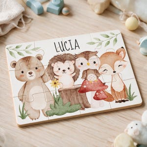 Könnte beinhalten: Ein rechteckiges Holzpuzzle mit einer Aquarellillustration eines Bären, eines Igels, einer Eule und eines Fuchses in einer Waldumgebung. Der Name LUCIA ist oben in schwarzen Großbuchstaben aufgedruckt und von grünen Blättern umgeben.